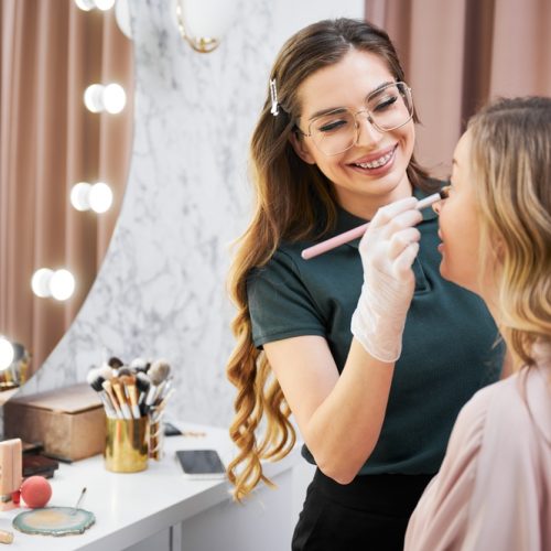 Smiling,Female,Stylist,In,Glasses,Applying,Foundation,On,Client,Face